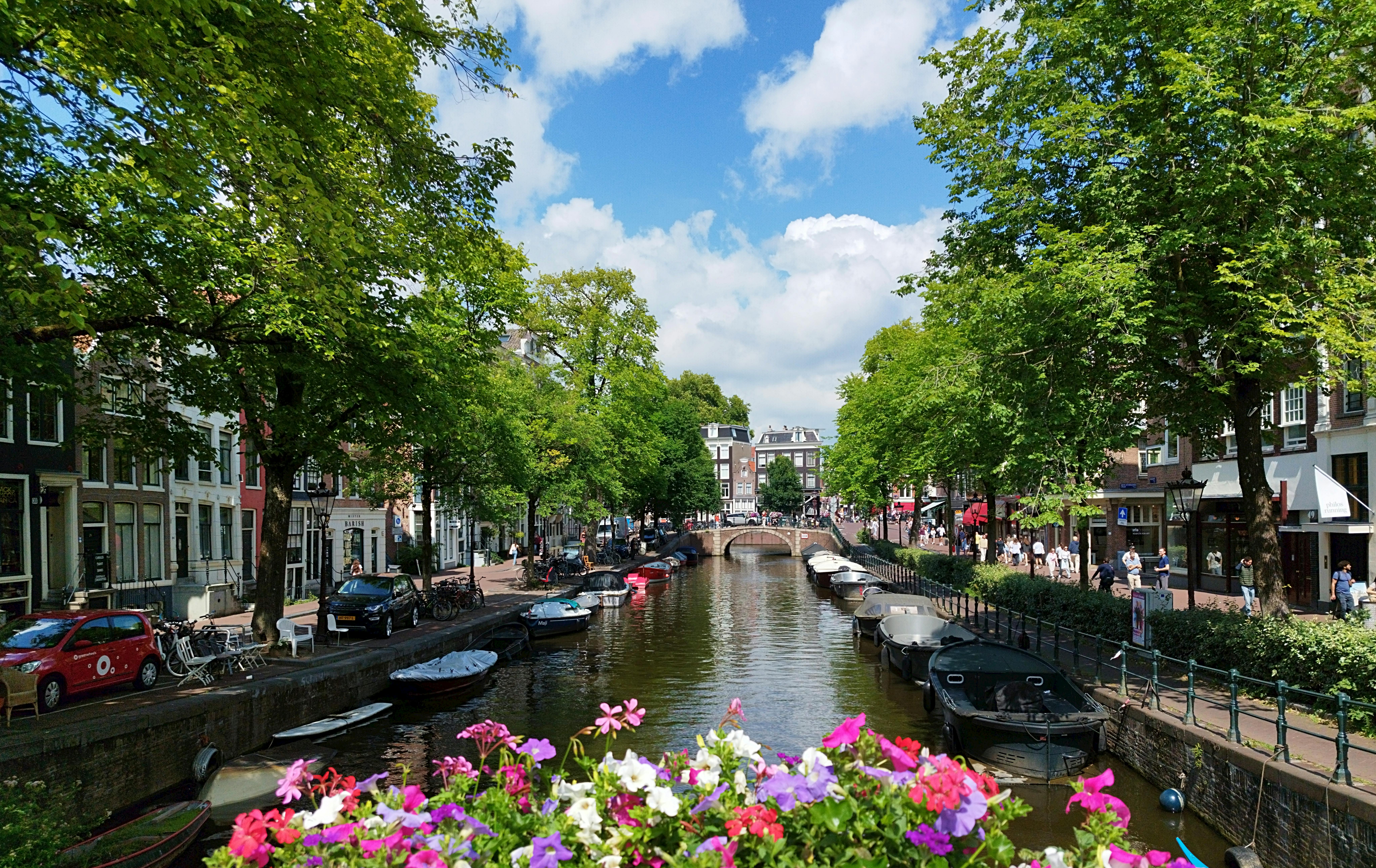 An Amsterdam canal lined with leafy trees, moored boats, and a stone bridge at the far end — the typical urban texture nomads come for.